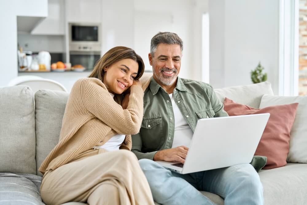 Happy middle aged couple using laptop relaxing on couch at home
